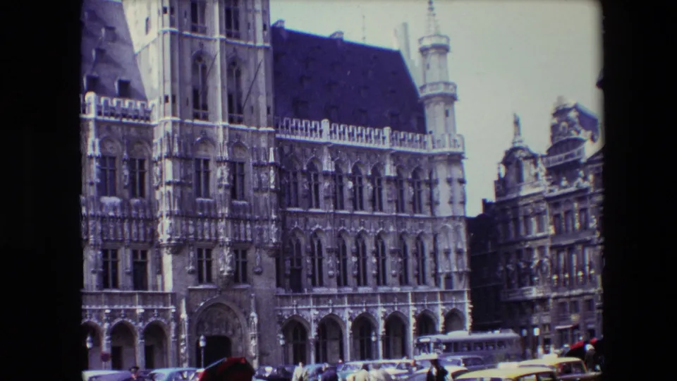 1967 Brussels home-movie still showing a closer crop of the Town Hall stonework and arched entrances, with pedestrians, parked cars, a bus, and a yellow taxi along the square.