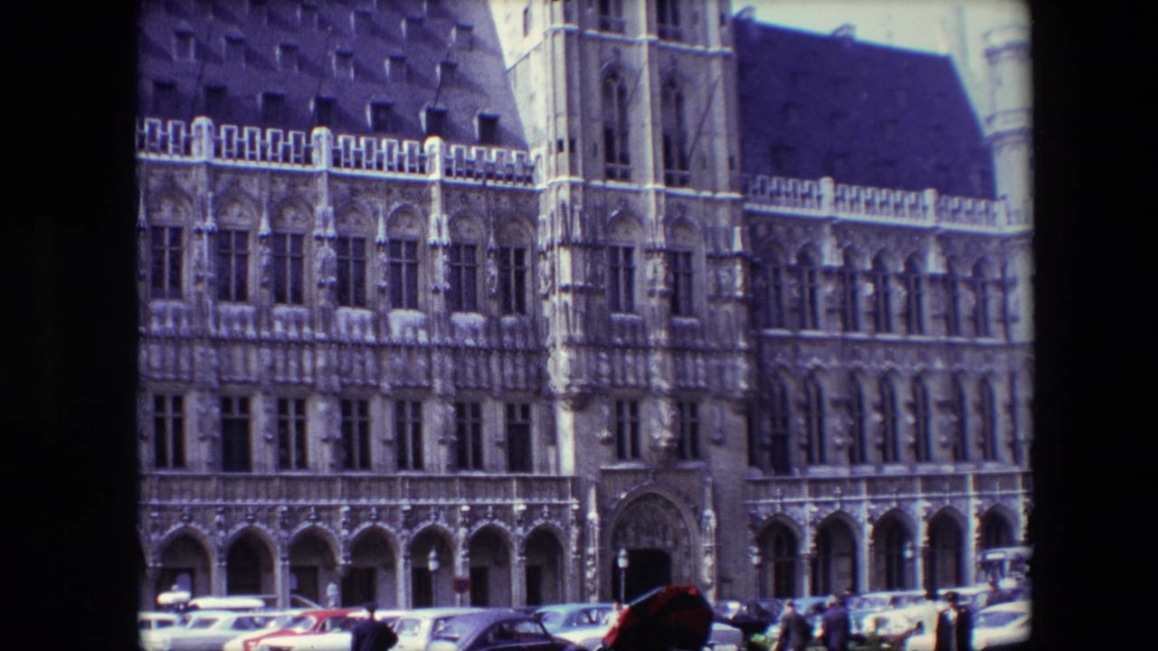 1967 Brussels frame with the camera on a detailed Gothic-style facade, including a tower bay, rows of windows, arched openings, parked cars, and distant pedestrians.