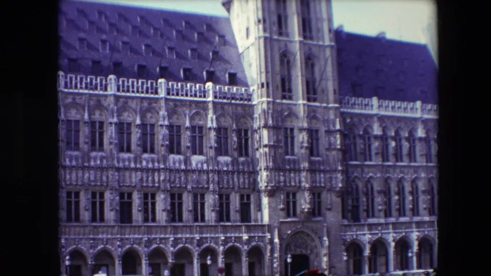 1967 Brussels 8mm frame showing a closer visitor view of the ornate stone facade, tower section, arched ground-floor bays, dark roof, and parked cars along the curb.