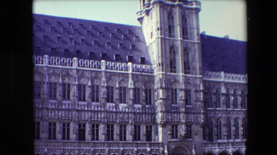 1967 Brussels frame holding on the historic Gothic-style facade, with a vertical tower section, decorative window bays, arched street-level openings, and parked cars in the foreground.
