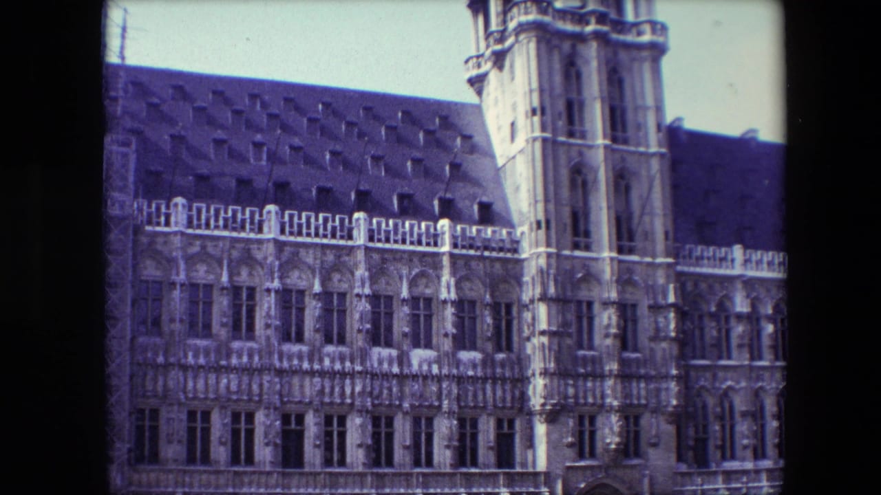 1967 Brussels home-movie frame showing an ornate Gothic-style masonry facade, dark roof, arched windows, and a tall tower under a pale sky.