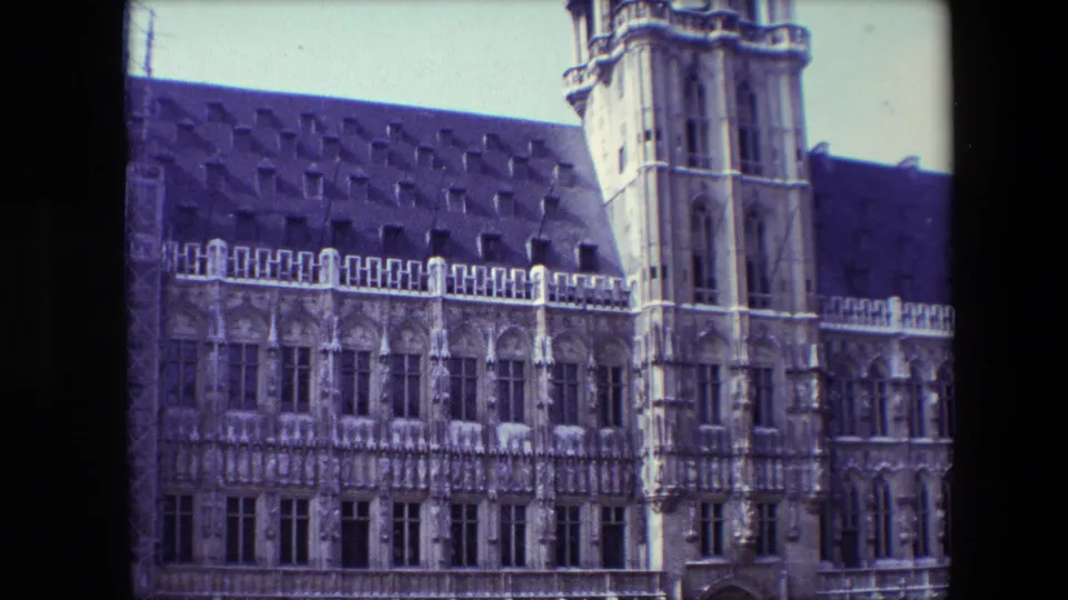 1967 Brussels home-movie frame showing an ornate Gothic-style masonry facade, dark roof, arched windows, and a tall tower under a pale sky.