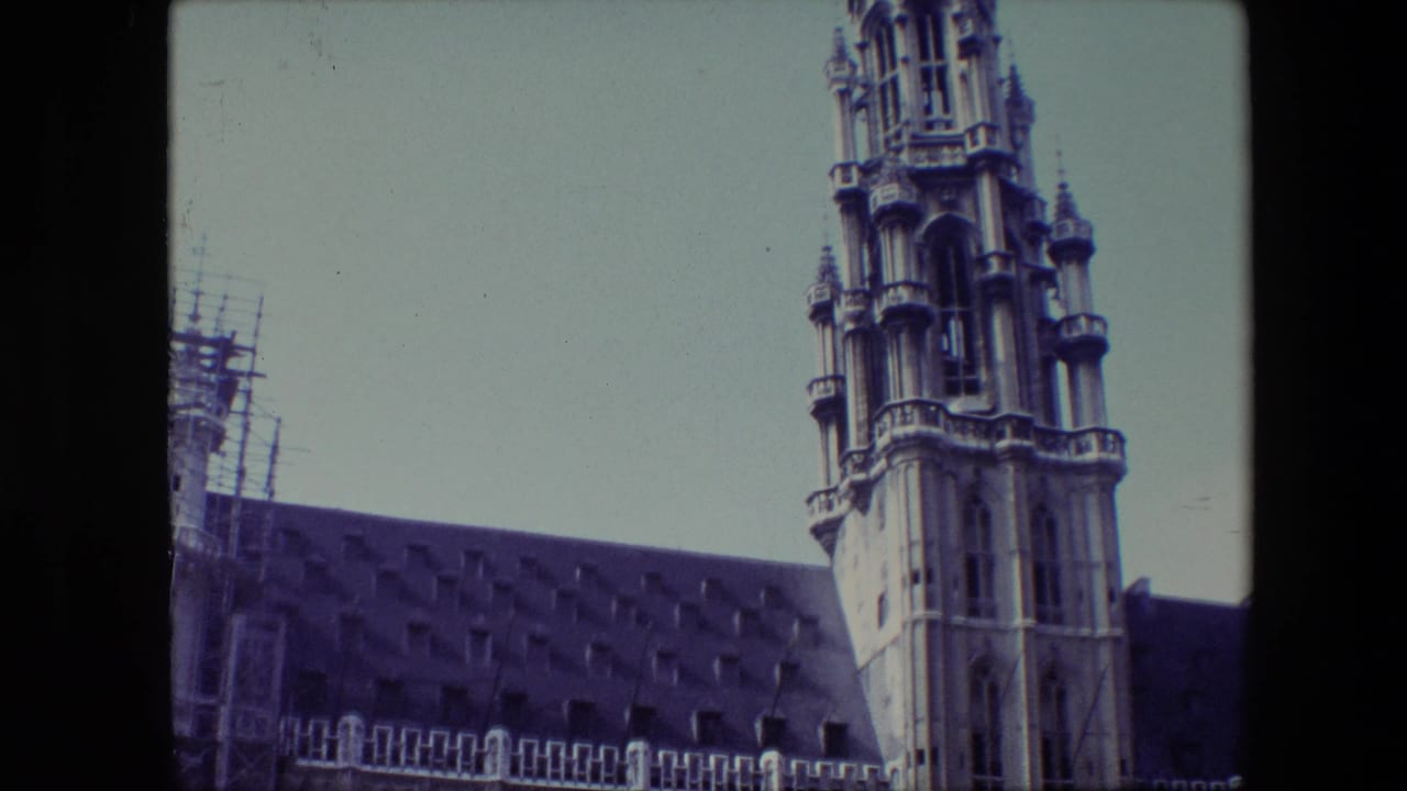 1967 Brussels home-movie frame showing an ornate Gothic cathedral spire above a dark roofline, with scaffolding visible along the left side.