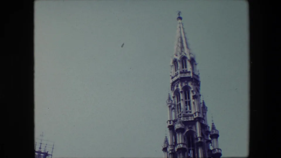Low-angle 1967 Brussels home-movie frame showing the upper portion of an ornate Gothic-style spire against a pale blue sky.