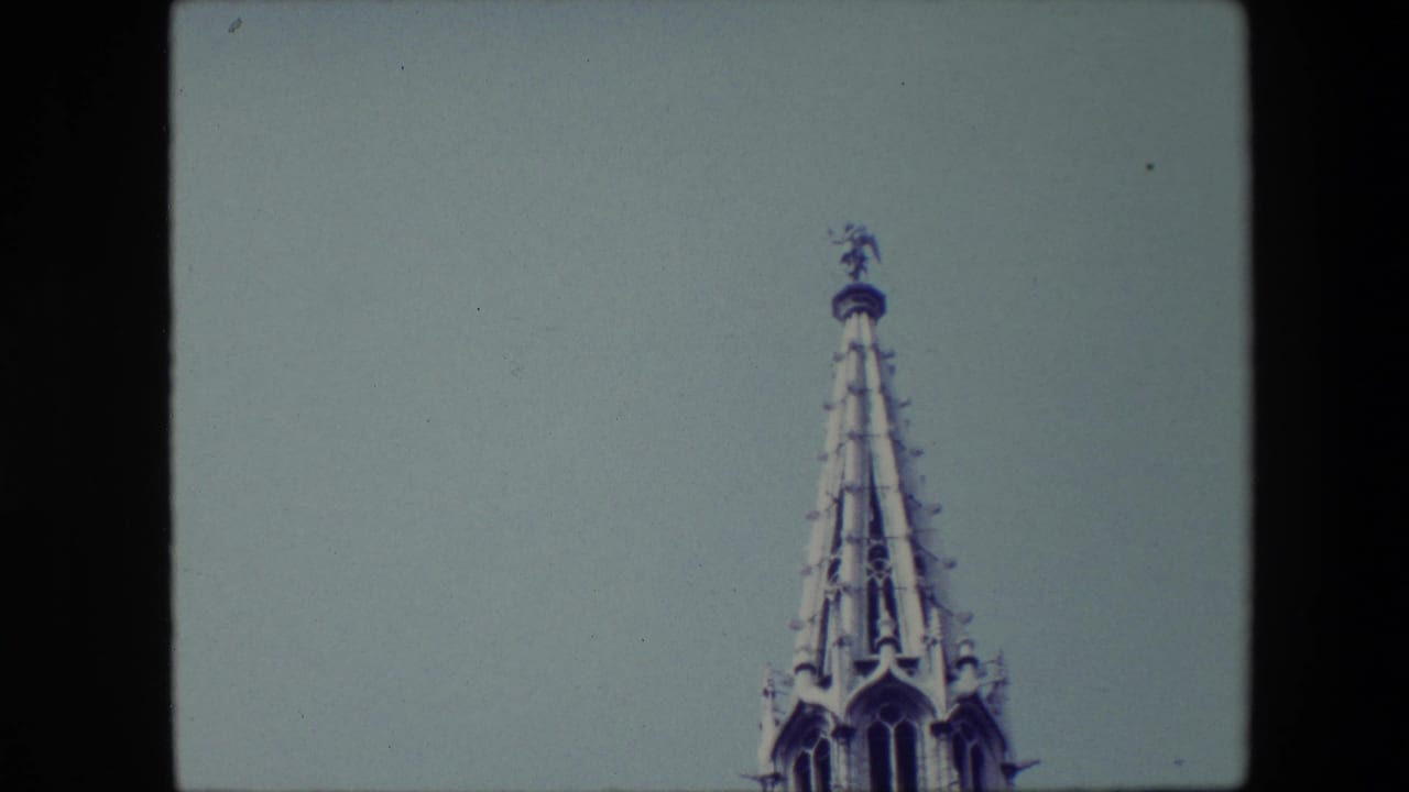 1967 Brussels home-movie frame showing the top of an ornate Gothic-style steeple with a small statue-like figure silhouetted against a pale blue sky.