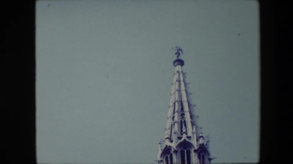 1967 Brussels home-movie frame showing the top of an ornate Gothic-style steeple with a small statue-like figure silhouetted against a pale blue sky.