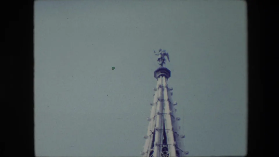1967 Brussels home-movie view of a tall Gothic-style spire detail with tracery and a small figure at the top, framed low against the sky.