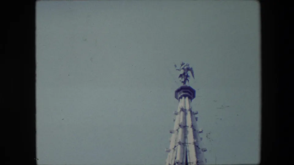 Final 1967 Brussels frame holding on the upper steeple and small statue-like figure, with visible 8mm softness and pale sky filling most of the view.
