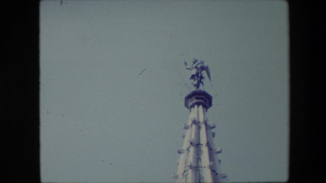 1967 Brussels home-movie frame showing the upper section of an ornate spire with a winged statue at the peak against a pale grey sky.
