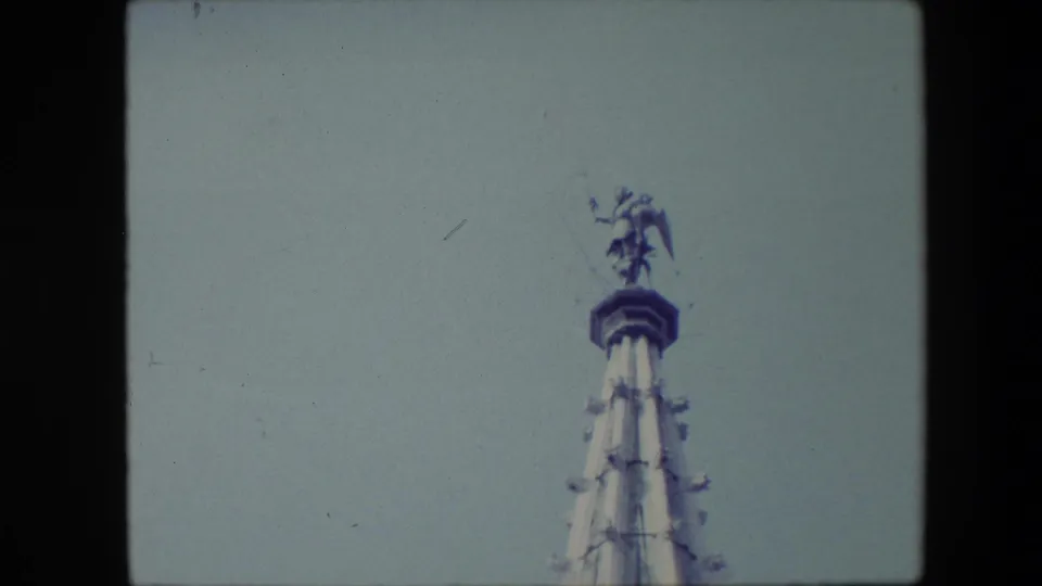 1967 Brussels home-movie frame showing the upper section of an ornate spire with a winged statue at the peak against a pale grey sky.
