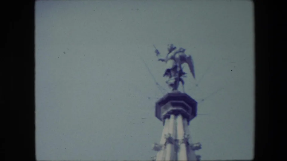 1967 Brussels frame with the camera held upward on the ornate spire top, bringing the winged statue and cap detail lower and larger in view.