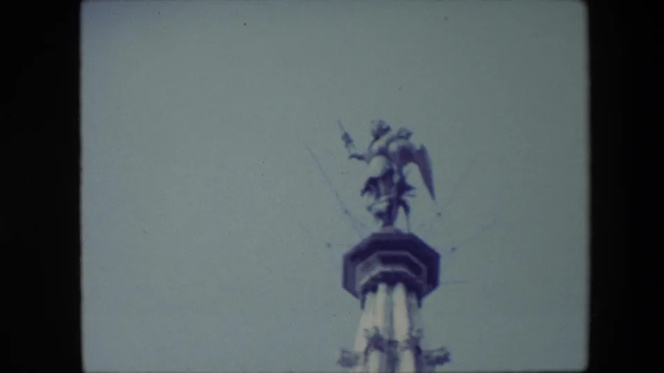 Final 1967 Brussels frame holding on the winged statue above the spire, with soft focus and blue-grey 8mm color visible across the sky.
