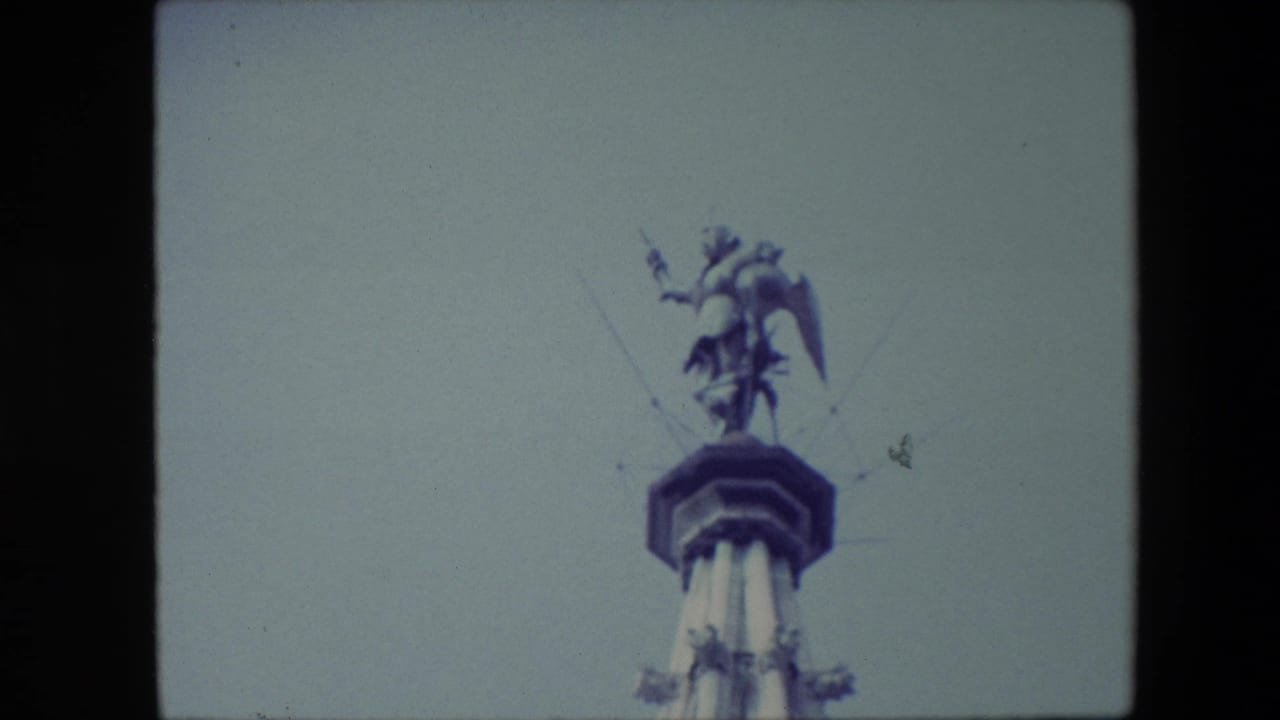 1967 Brussels 8mm frame showing a low-angle view of a winged male statue with a spear standing on a tall monument against a pale blue sky.