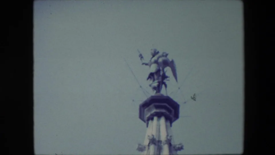 1967 Brussels 8mm frame showing a low-angle view of a winged male statue with a spear standing on a tall monument against a pale blue sky.
