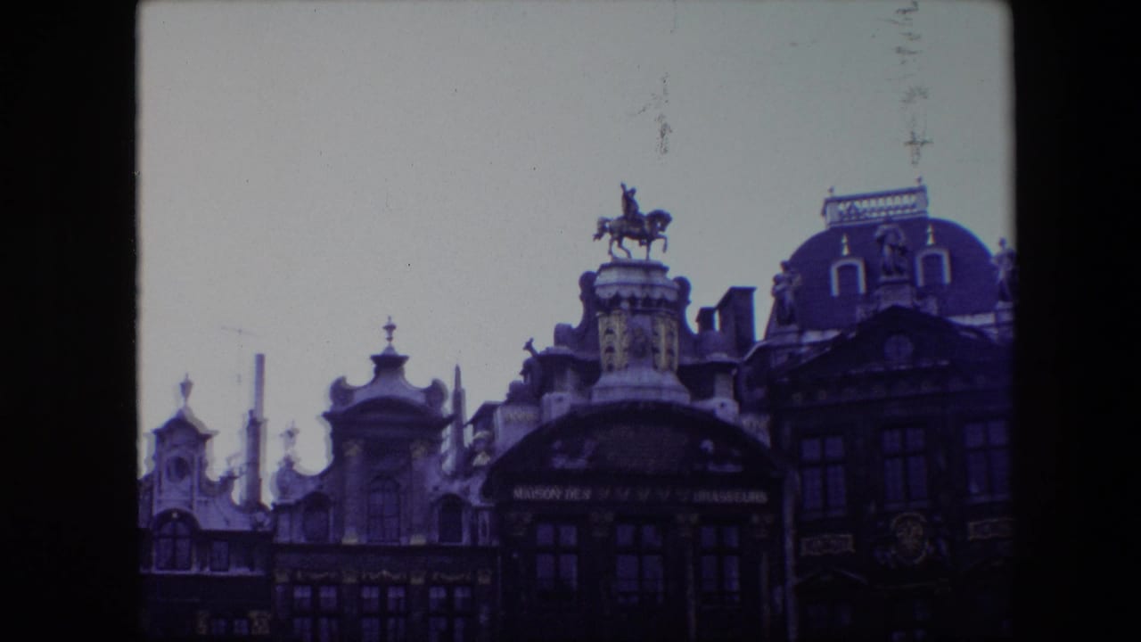 1967 8mm frame of ornate Brussels Grand Place architecture with an equestrian statue mounted above a historic facade under an overcast sky.