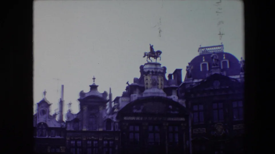 1967 8mm frame of ornate Brussels Grand Place architecture with an equestrian statue mounted above a historic facade under an overcast sky.