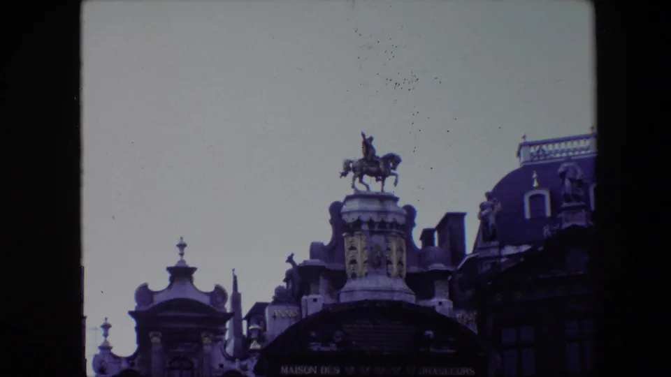The camera frames the rooftop equestrian statue and adjacent ornate Grand Place rooflines in Brussels, with soft 8mm color and visible film border.