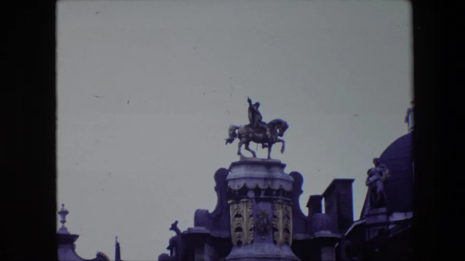 A tighter 1967 Brussels home-movie frame shows the equestrian rooftop statue above carved facade elements, with neighboring roof details at the edges.