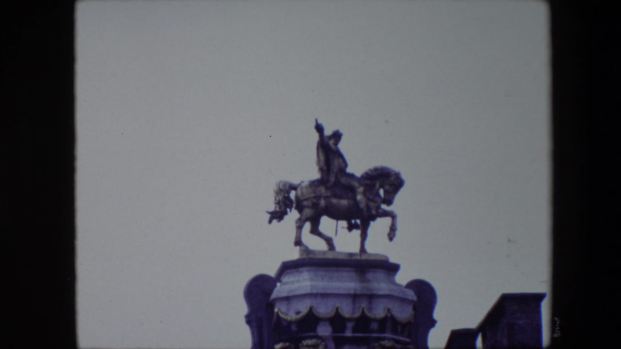 1967 Brussels home-movie frame showing a distant bronze equestrian statue on a high pedestal, with the rider raising one arm against a pale sky.