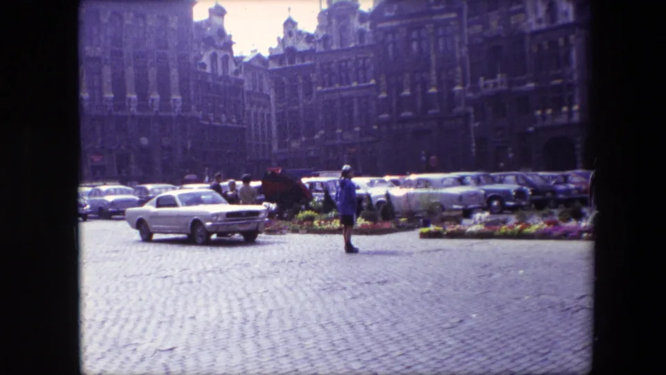 A Brussels historic square in 1967 shows cobblestones, parked 1960s cars, small pedestrian figures, flower beds, and ornate building facades in soft 8mm color.