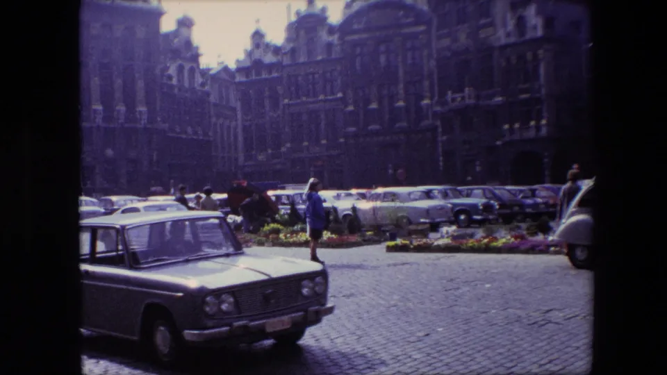 A classic 1960s car occupies the foreground of a Brussels square, with pedestrians, flower beds, parked vehicles, cobblestones, and historic architecture behind it.