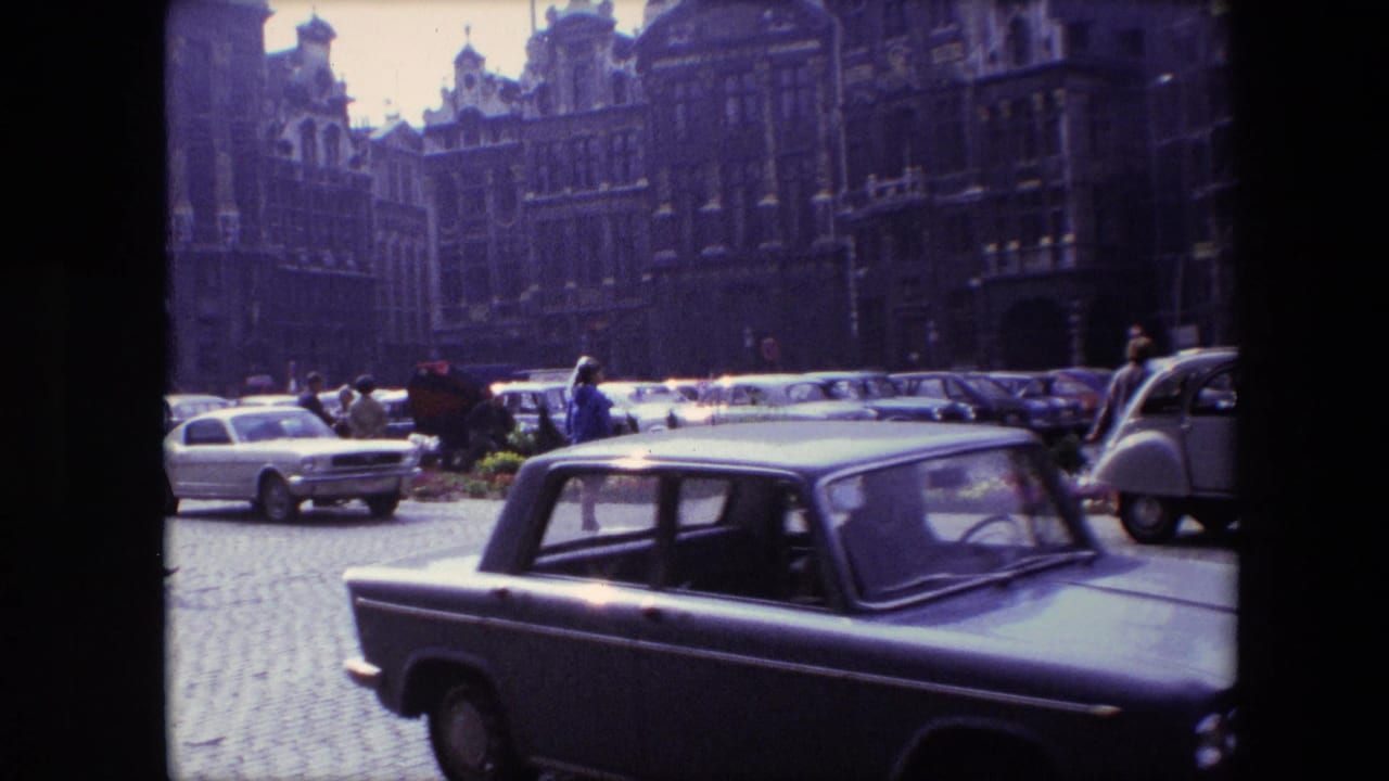 1967 Brussels home-movie frame showing a dark car moving across a cobblestone square, with parked mid-century cars and ornate historic facades in the background.