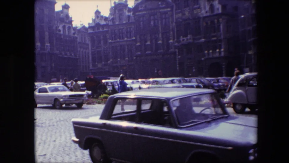 1967 Brussels home-movie frame showing a dark car moving across a cobblestone square, with parked mid-century cars and ornate historic facades in the background.