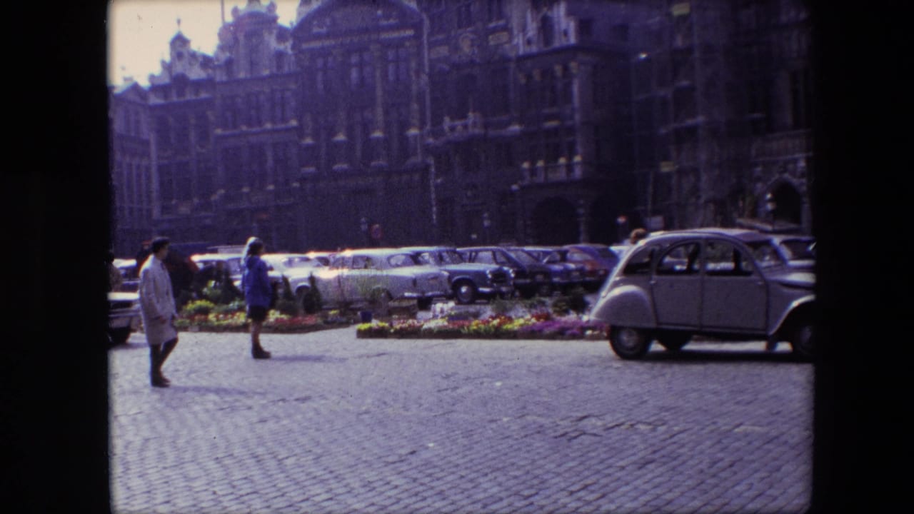 1967 Brussels frame with pedestrians crossing a cobblestone square beside flower beds, rows of parked cars, and a light-colored compact European car.