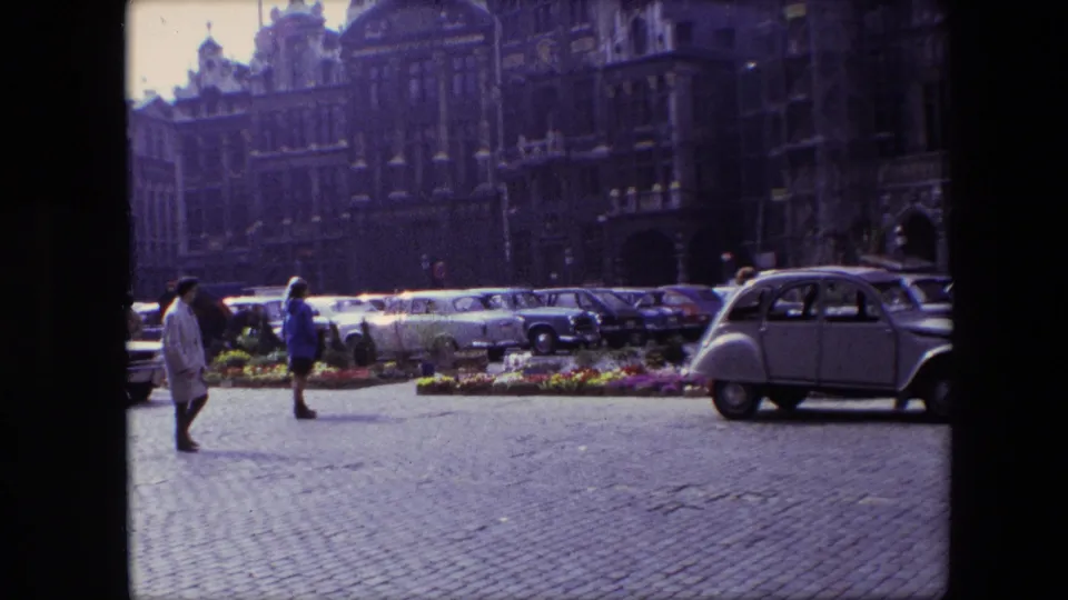 1967 Brussels frame with pedestrians crossing a cobblestone square beside flower beds, rows of parked cars, and a light-colored compact European car.