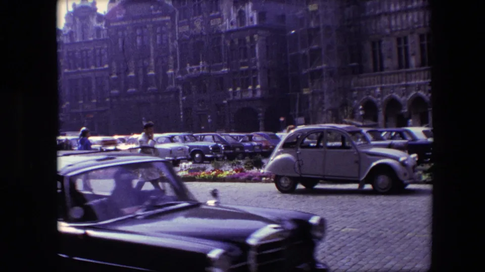 1967 Brussels home-movie frame showing a dark car crossing the foreground while a light-colored compact car, pedestrians, parked cars, and historic facades remain behind it.