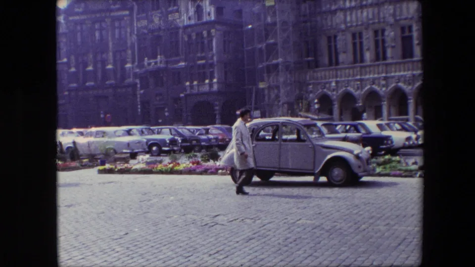 1967 Brussels frame showing a man in a light coat crossing in front of a light-colored compact European car, with parked cars and historic buildings behind him.