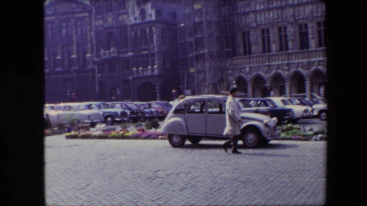 1967 Brussels home-movie frame showing a cobblestone square with compact parked cars, a pedestrian in a pale coat, flower displays, and a historic arched facade.
