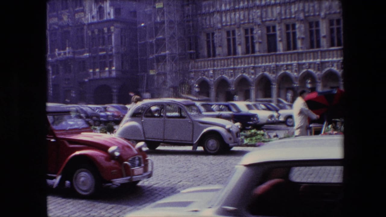 1967 Brussels frame with a red compact car moving across the foreground, parked cars behind it, flower displays, and historic stone arches in the background.