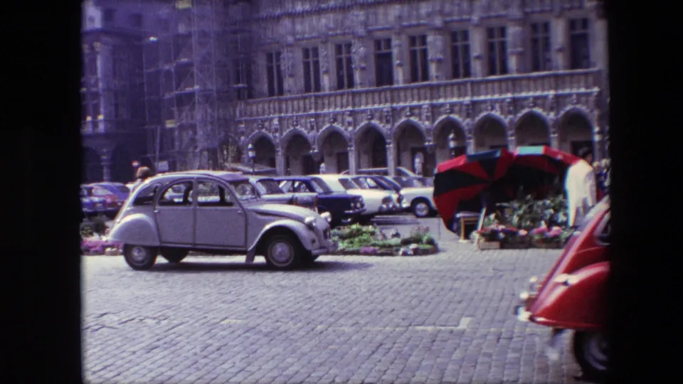 1967 Brussels home-movie still showing a parked compact car, flower displays, red-and-dark striped umbrellas, cobblestones, and a historic facade with arched openings.