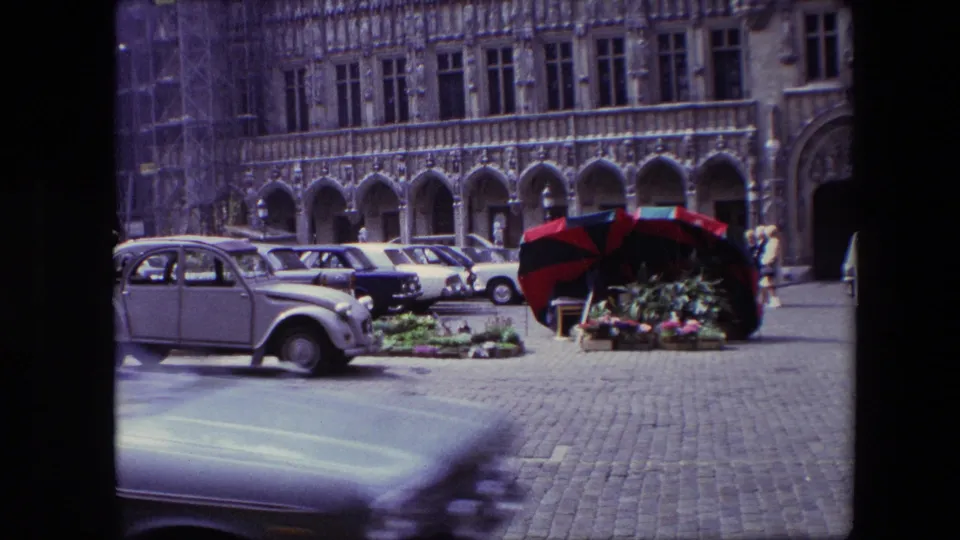 1967 Brussels frame with motion blur from a passing foreground car, a parked compact car, flower displays, striped umbrellas, and arched historic architecture beyond.