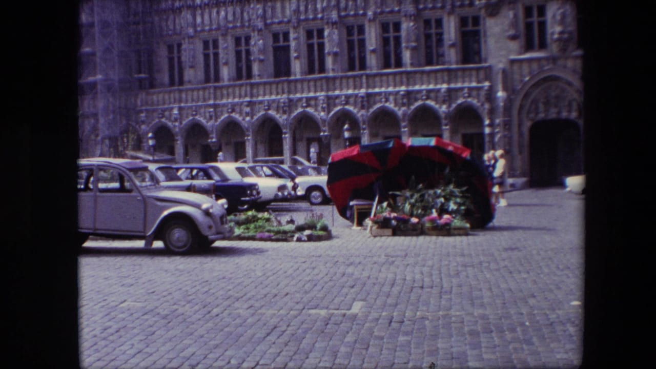 1967 Brussels home-movie frame showing a cobblestone square, parked mid-century cars, a red-and-black flower stall umbrella, and an ornate arched stone facade.