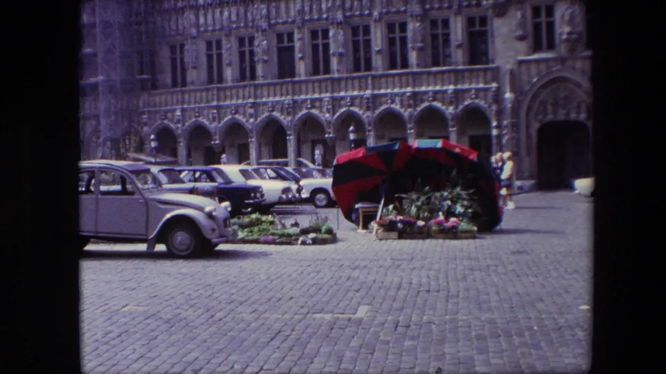 1967 Brussels home-movie frame showing a cobblestone square, parked mid-century cars, a red-and-black flower stall umbrella, and an ornate arched stone facade.
