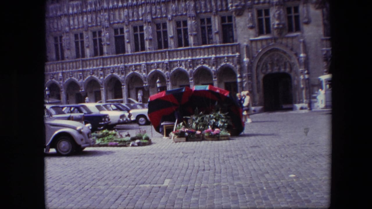 1967 Brussels street view with a red-and-black umbrella over flowers, compact cars parked along the square, and pedestrians near an elaborate arched facade.