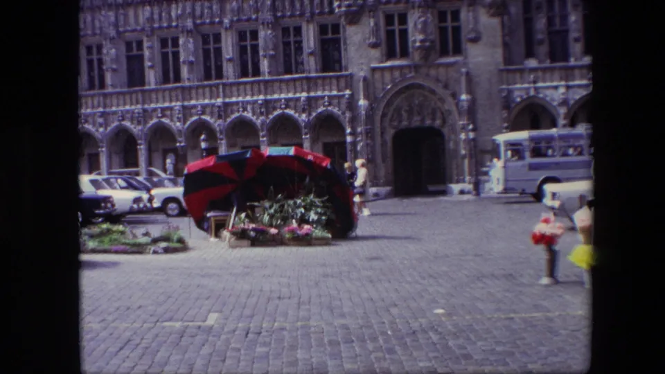 1967 Brussels frame with a white bus passing the ornate stone building, a flower stall under a striped umbrella, parked cars, and pedestrians at the edge of the square.