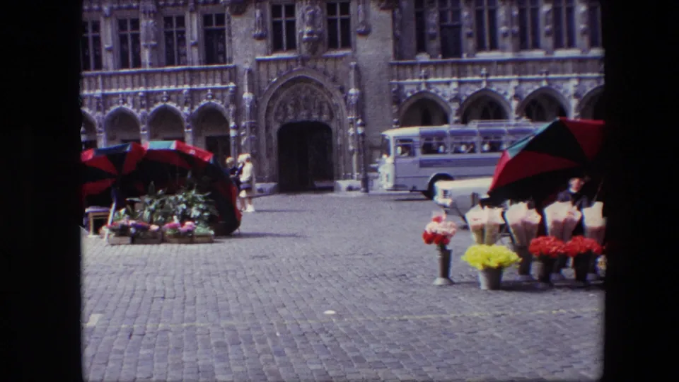 1967 Brussels home-movie frame showing a white bus, buckets of bright flowers, a red-and-black vendor umbrella, cobblestones, and the arched stone building behind.