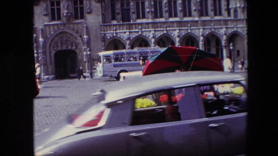 A classic car moves across the foreground, partly obscuring the Brussels flower stall, umbrellas, bus, pedestrians, and carved stone building behind it.