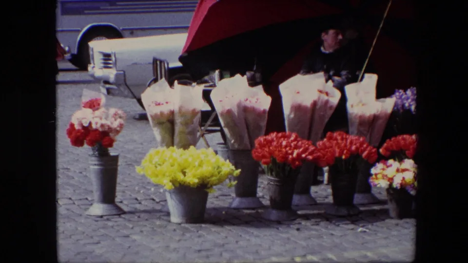 A closer 1967 Brussels view shows bouquets arranged in metal cans beneath a red and black umbrella, with the vendor partly visible and a vehicle passing behind the stall.