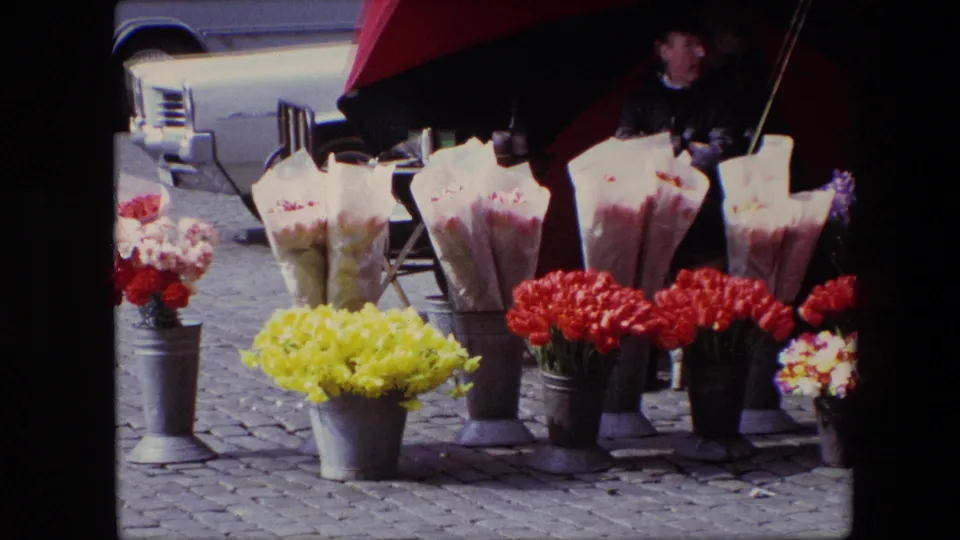 The final 1967 Brussels frame concentrates on the flower stall, with yellow and red bouquets in metal cans, wrapped flowers, cobblestone paving, and the vendor partly visible beneath the umbrella.