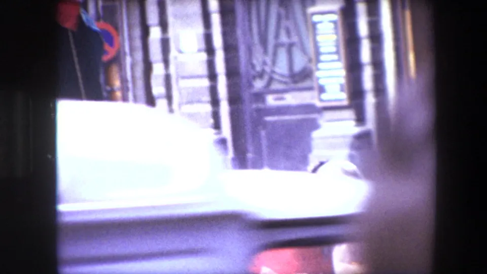 The final 1967 Brussels frame shifts away from the flowers toward a blurred street facade and a passing white vehicle, retaining the obstructed home-movie framing.