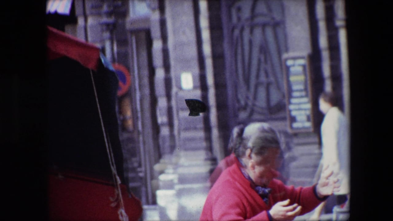 In Brussels home-movie footage from 1967, a gray-haired woman in a red coat appears to handle items at a street stall beneath a red-and-black umbrella, with a stone building and passerby behind her.