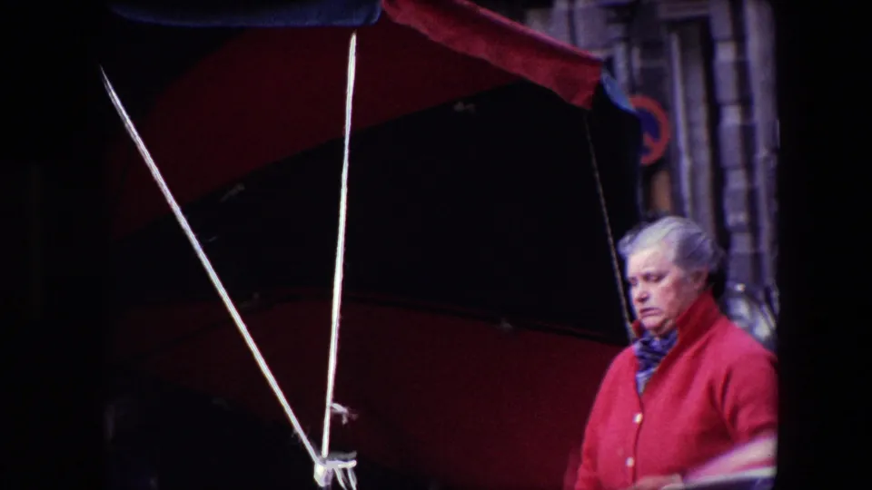 A gray-haired woman in a red coat stands under the red-and-black market umbrella in the Brussels 1967 footage, with the stall area partly shaded and the stone street backdrop still visible.