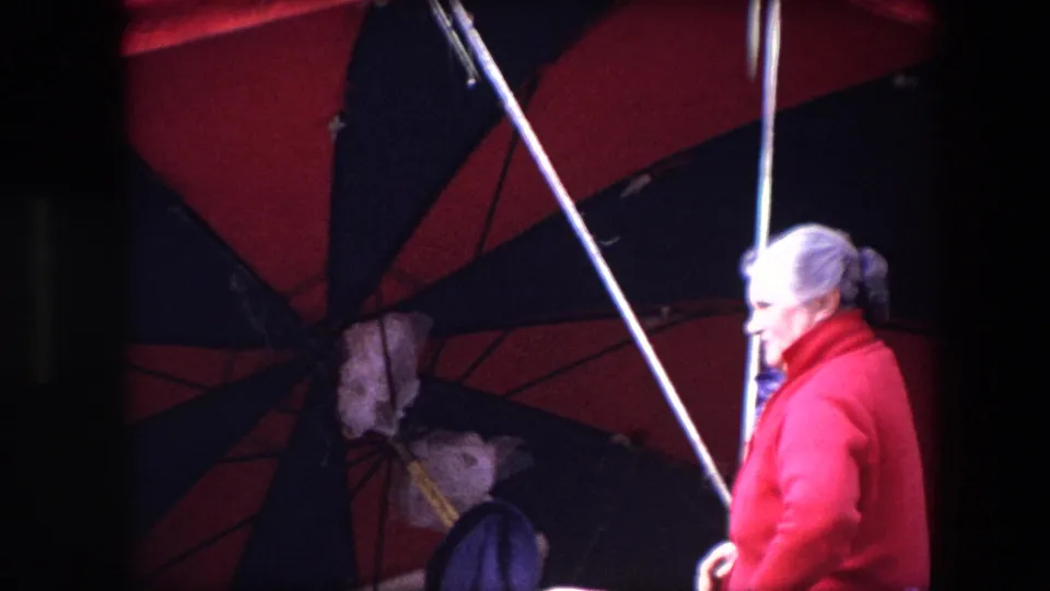 The final frame shows the woman in profile beside the large red-and-black umbrella, her red coat catching bright light while the market stall and street setting remain partially obscured by shade.