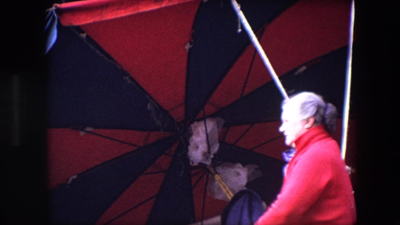 In Brussels 1967 home-movie footage, an older woman in a bright red sweater is seen in profile beneath a large red and dark umbrella near a dim building facade.