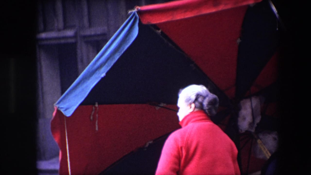 Older woman in red is seen from behind beside a red and dark umbrella.