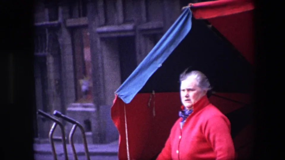 In the Brussels 1967 8mm frame, the older woman in a red sweater faces the camera with a serious or concerned expression while standing beside the large red and dark umbrella.